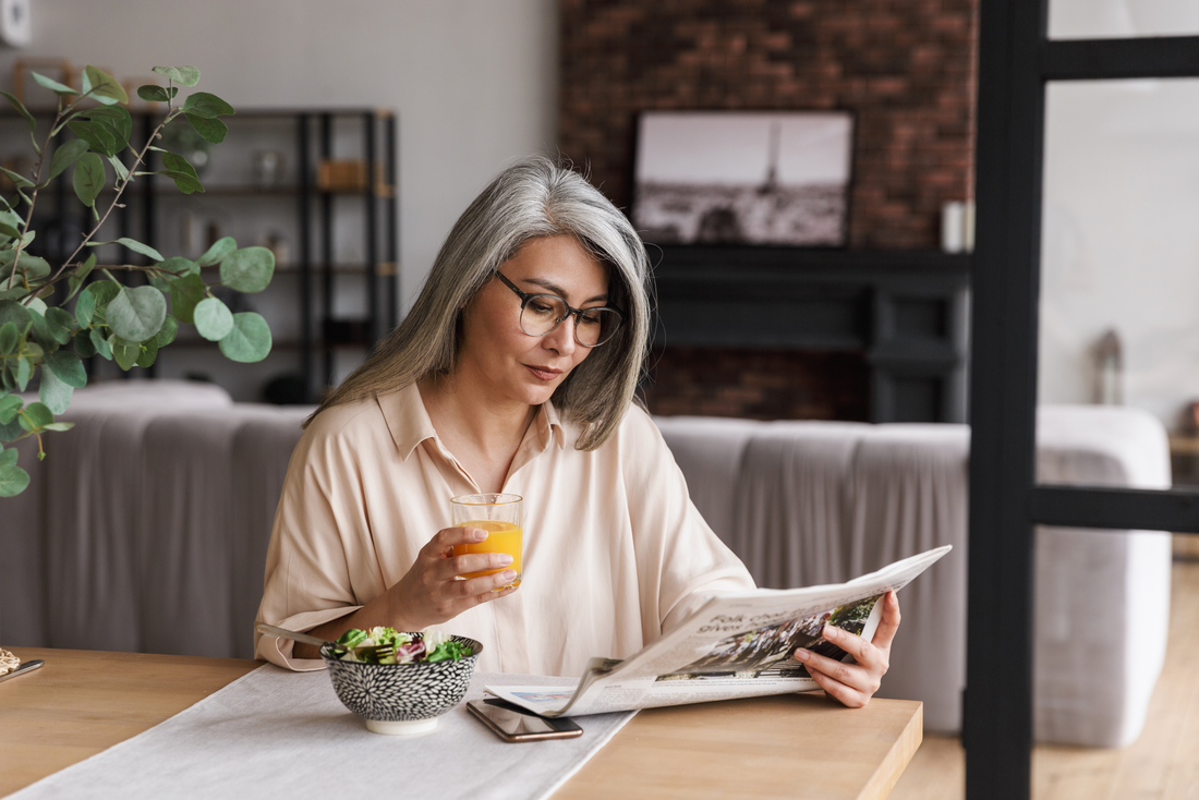 Eine Frau liest eine Zeitung am Tisch, neben einer Schüssel mit Obst und einem Glas Orangensaft.
